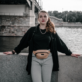A woman with brown hair wearing a black top, black jacket, grey jeans, and a tan fanny pack. 